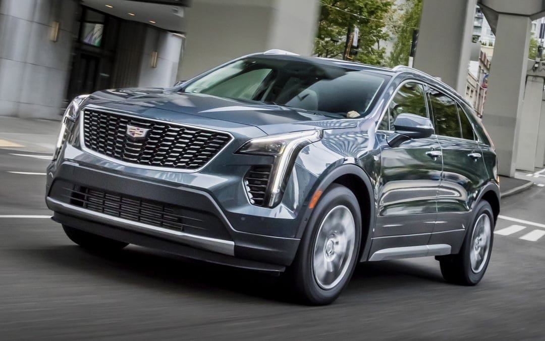 Cadillac SUV driving on a city street with motion blurred wheels against modern architecture and trees.