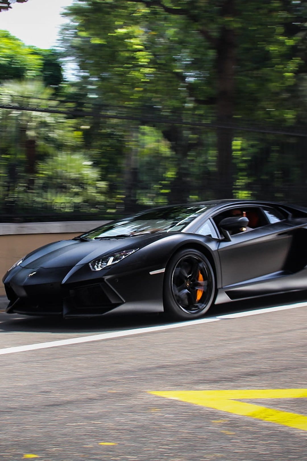 Matte black Lamborghini Aventador with orange calipers speeding past green trees on a paved race track.