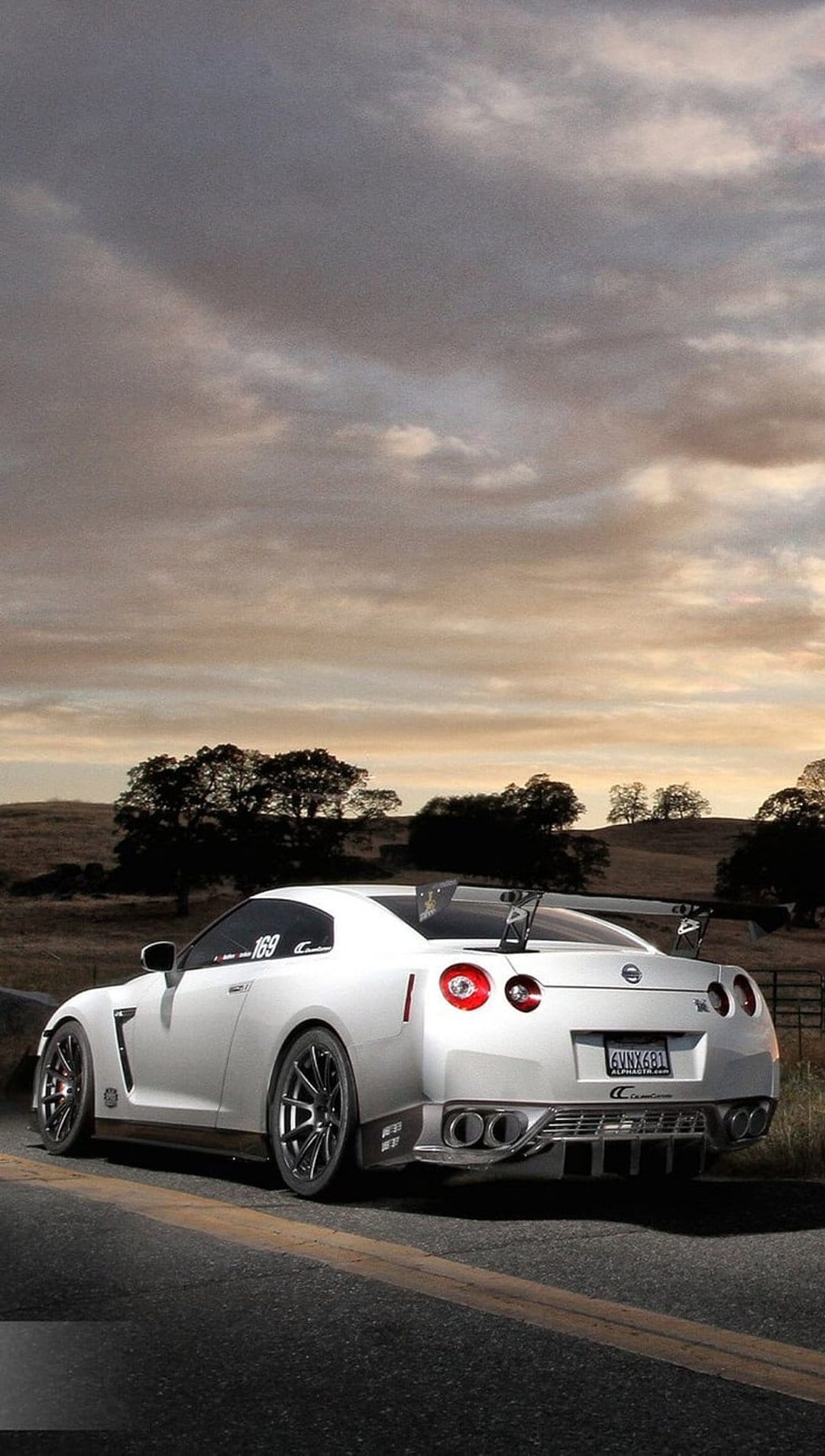 White Nissan GT-R R35 with a large rear spoiler parked on a rural asphalt road under a dramatic cloudy sky.