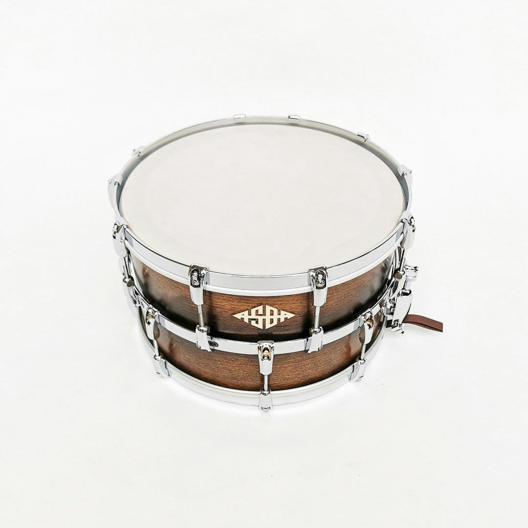 Wooden snare drum with polished silver lugs and a white drumhead positioned against a bright studio background.