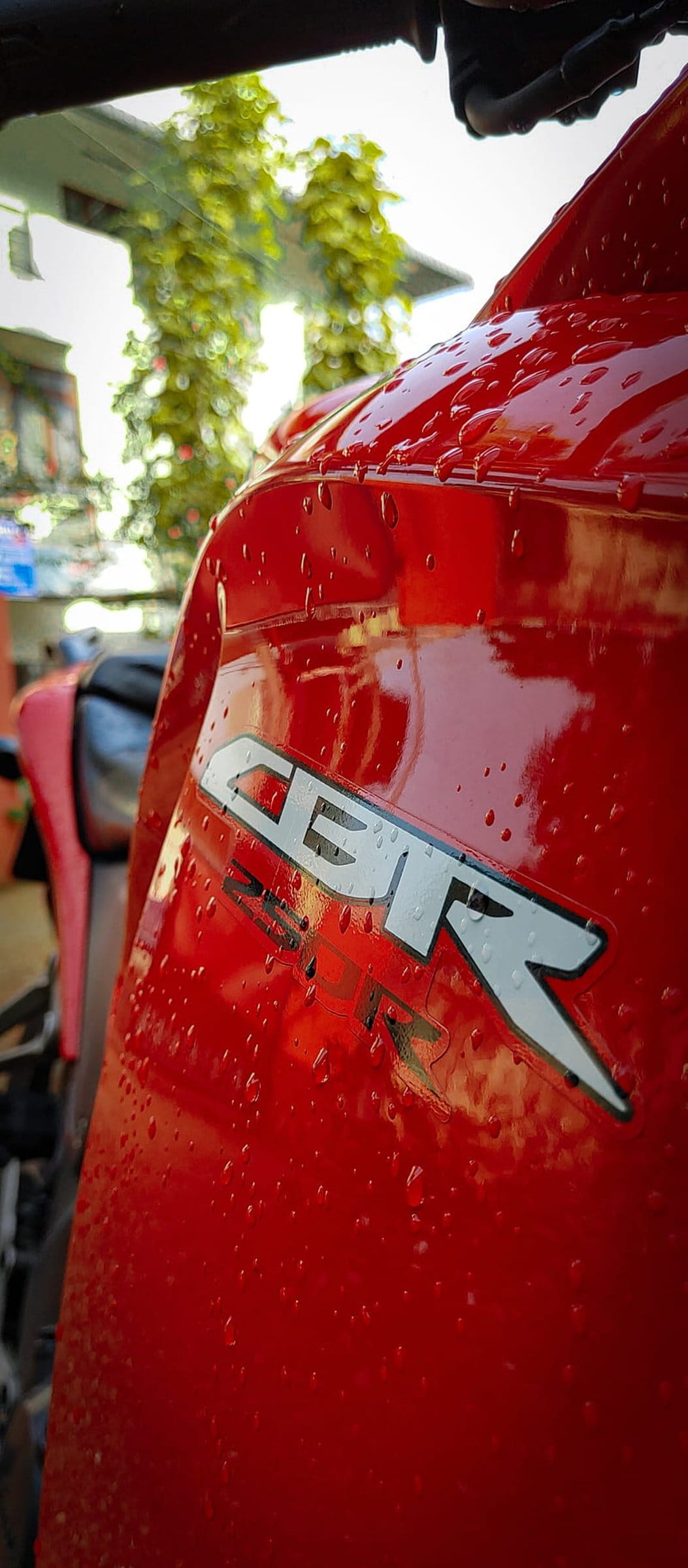 Red Honda CBR motorcycle fairing with silver logo and water droplets against a blurred green background.