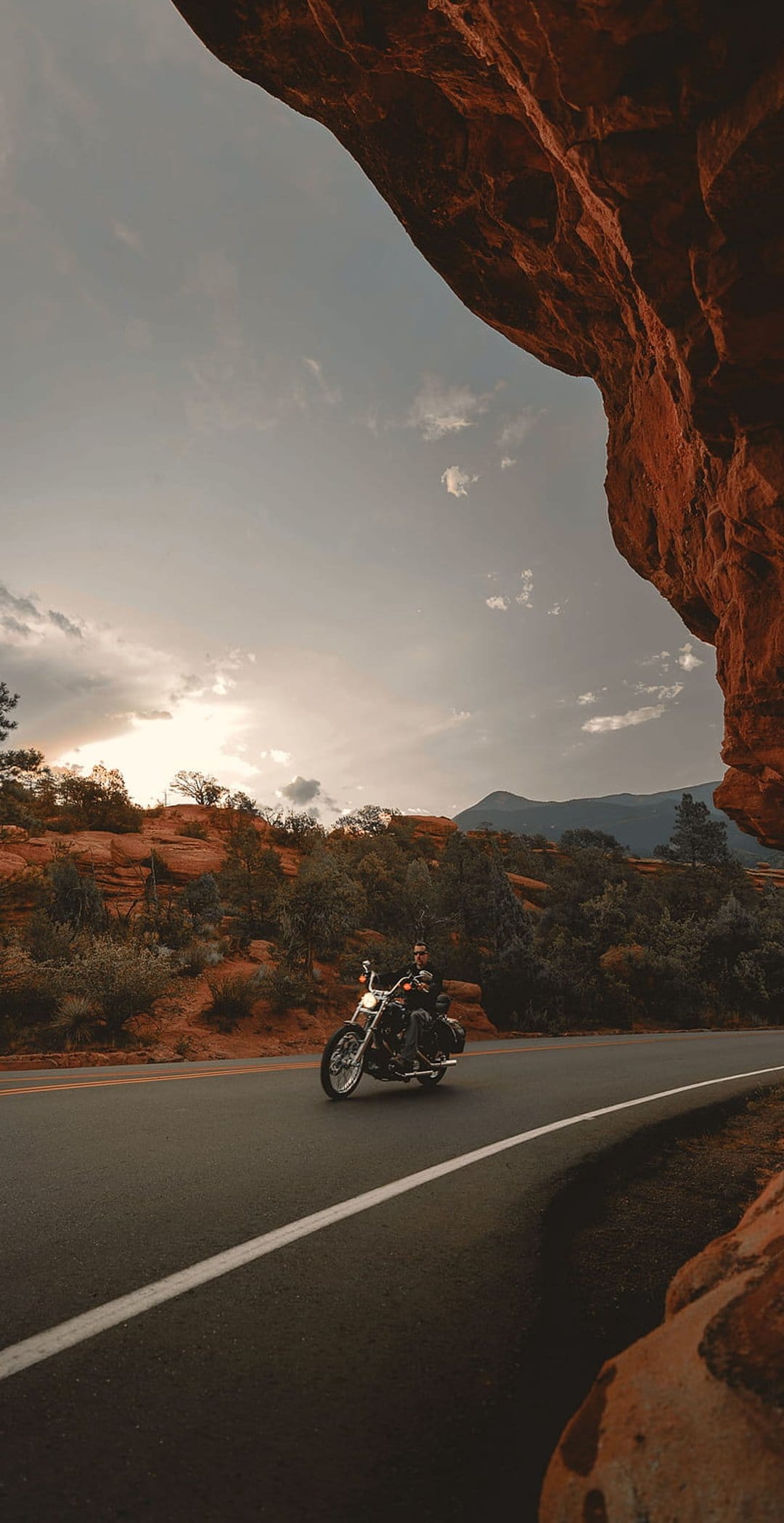 Lone motorcyclist rides a black cruiser down a winding desert road surrounded by towering red rock formations.