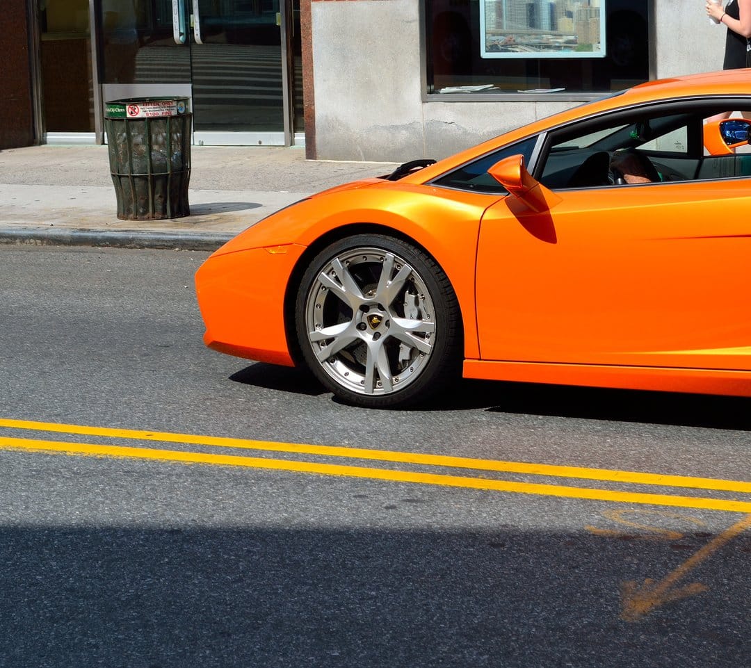 Orange Lamborghini supercar speeding down a sunny urban road with silver wheels and sharp shadows visible.