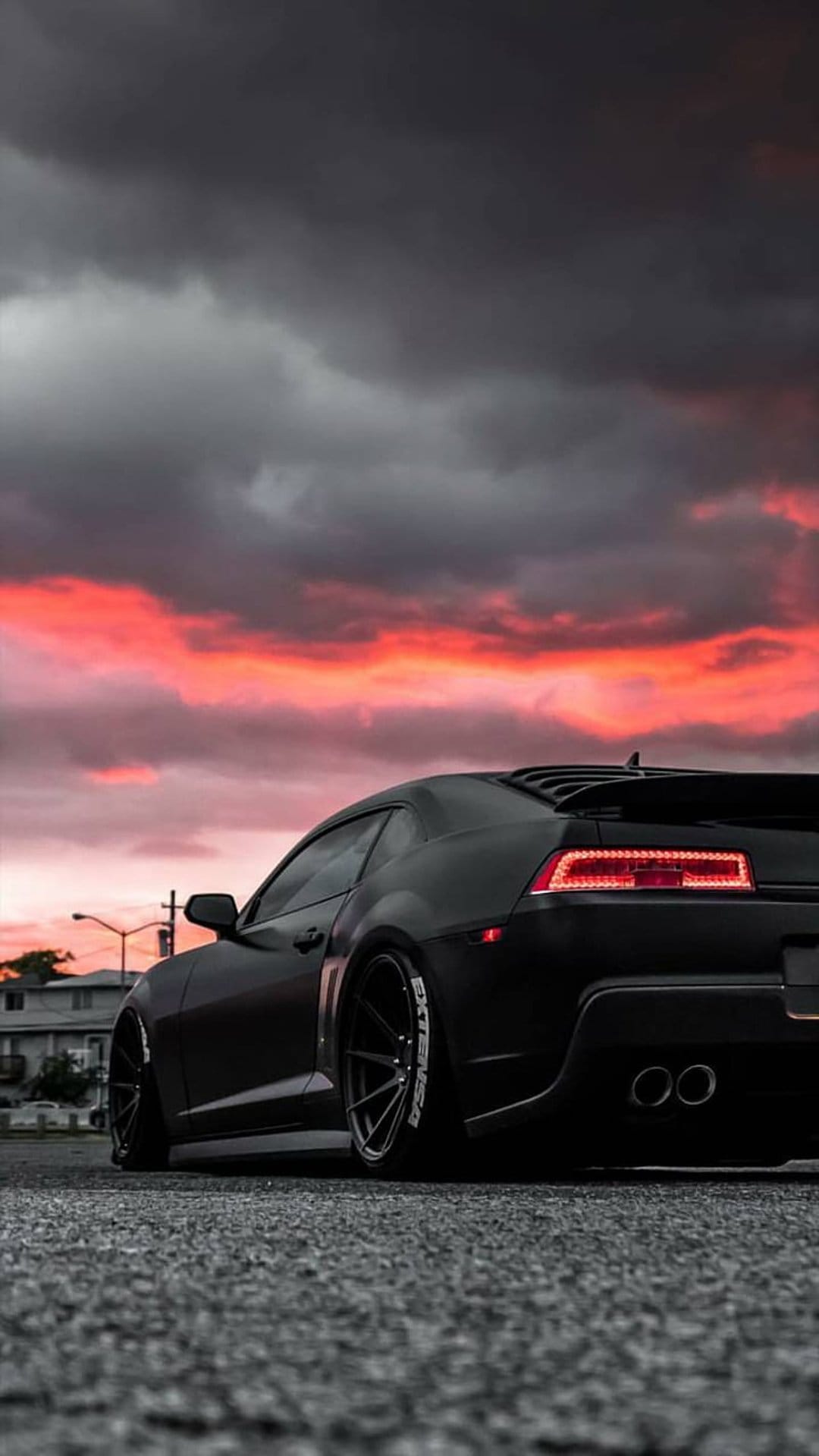 Black muscle car showing rear taillights on an asphalt road under a dramatic orange and red sunset sky.
