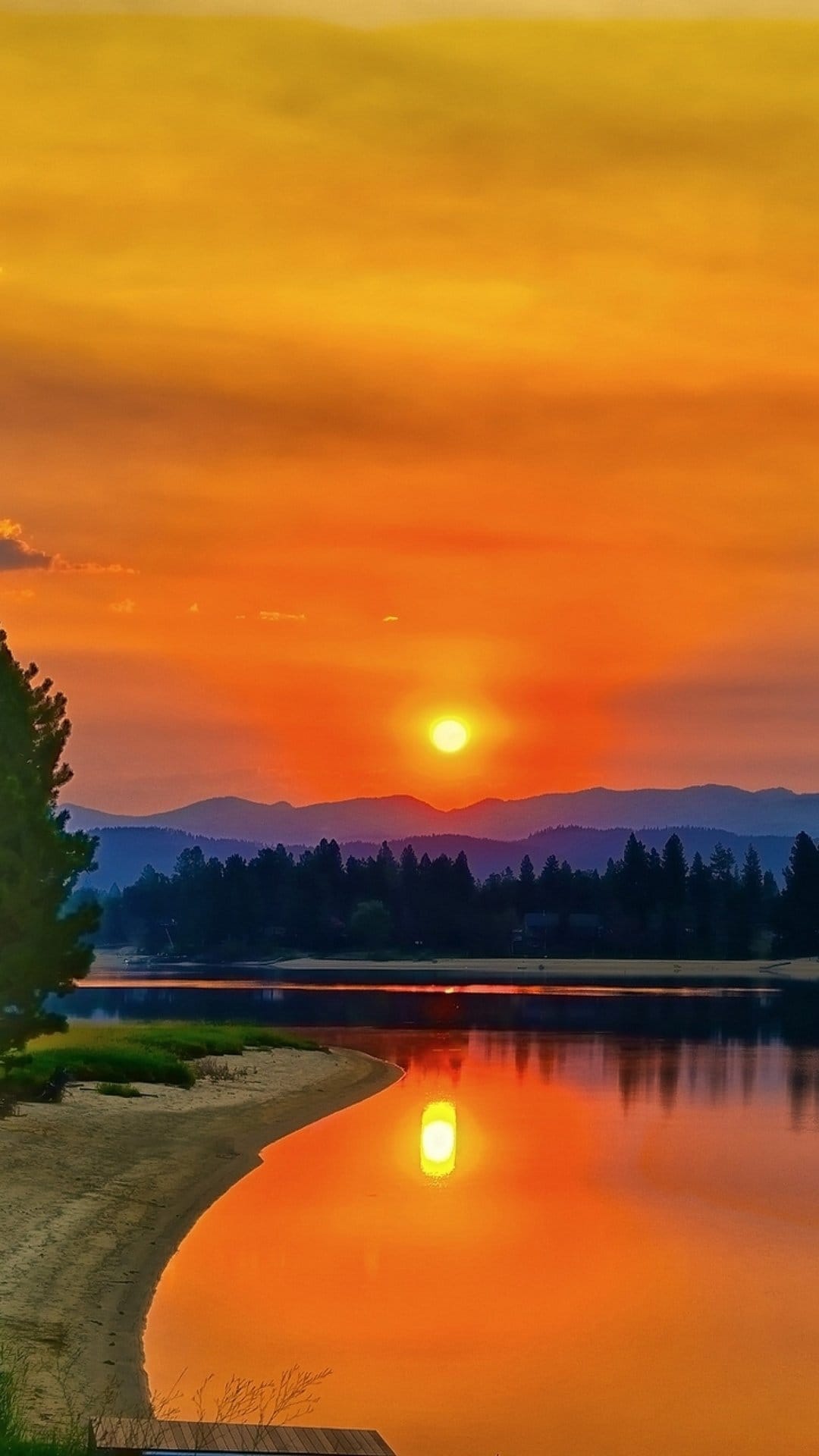 Intense red sunset reflecting in a still lake with mountain silhouettes and a sandy beach in the foreground.