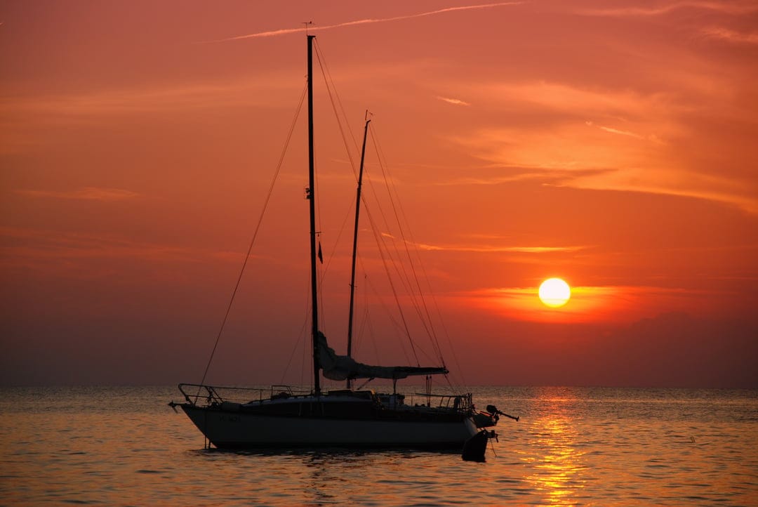 Sailboat silhouette rests on calm ocean waters under a vibrant orange sunset with glowing sun on the horizon.