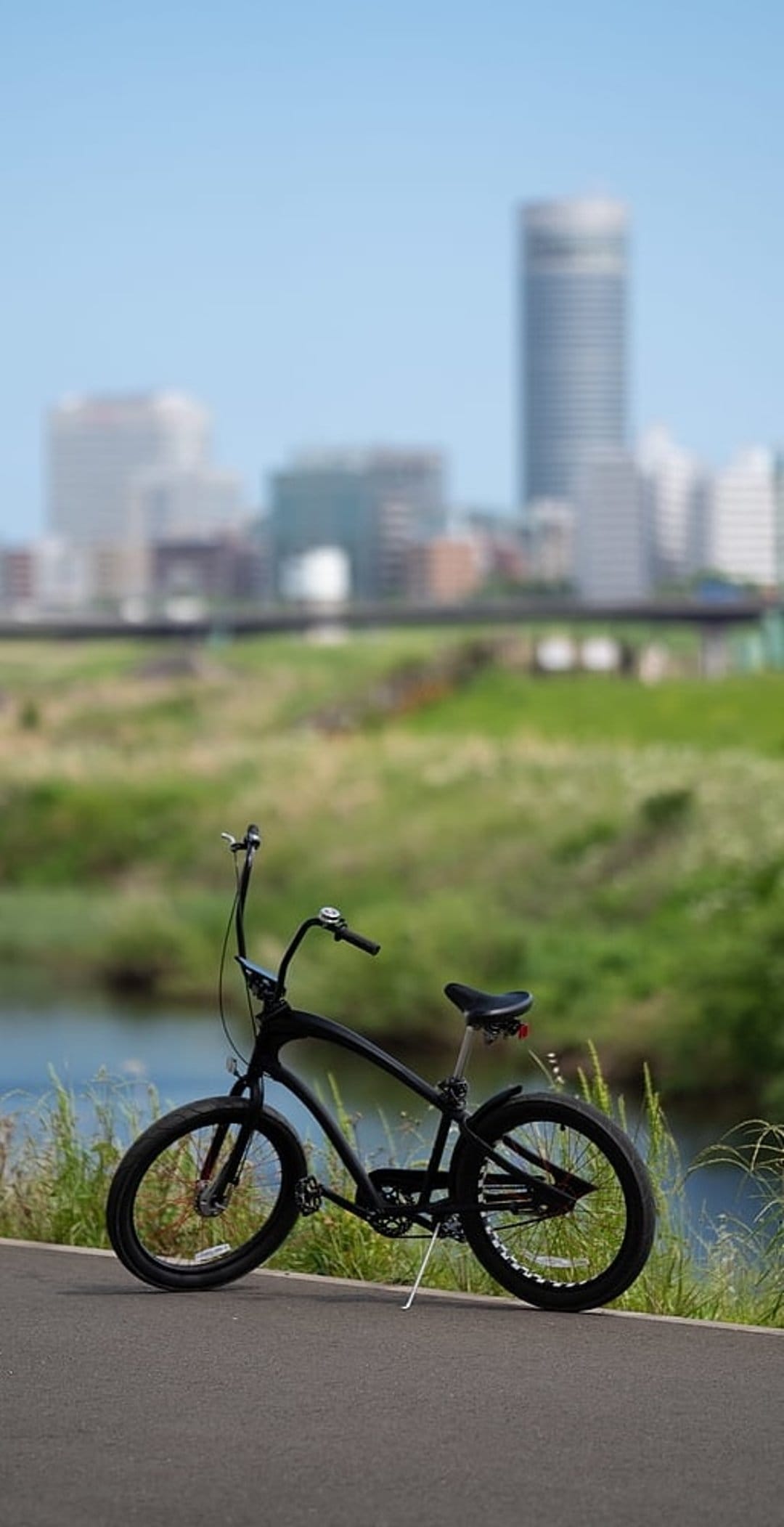 Black cruiser bicycle stands on a paved trail beside a river with a blurred city skyline and bridge behind it.