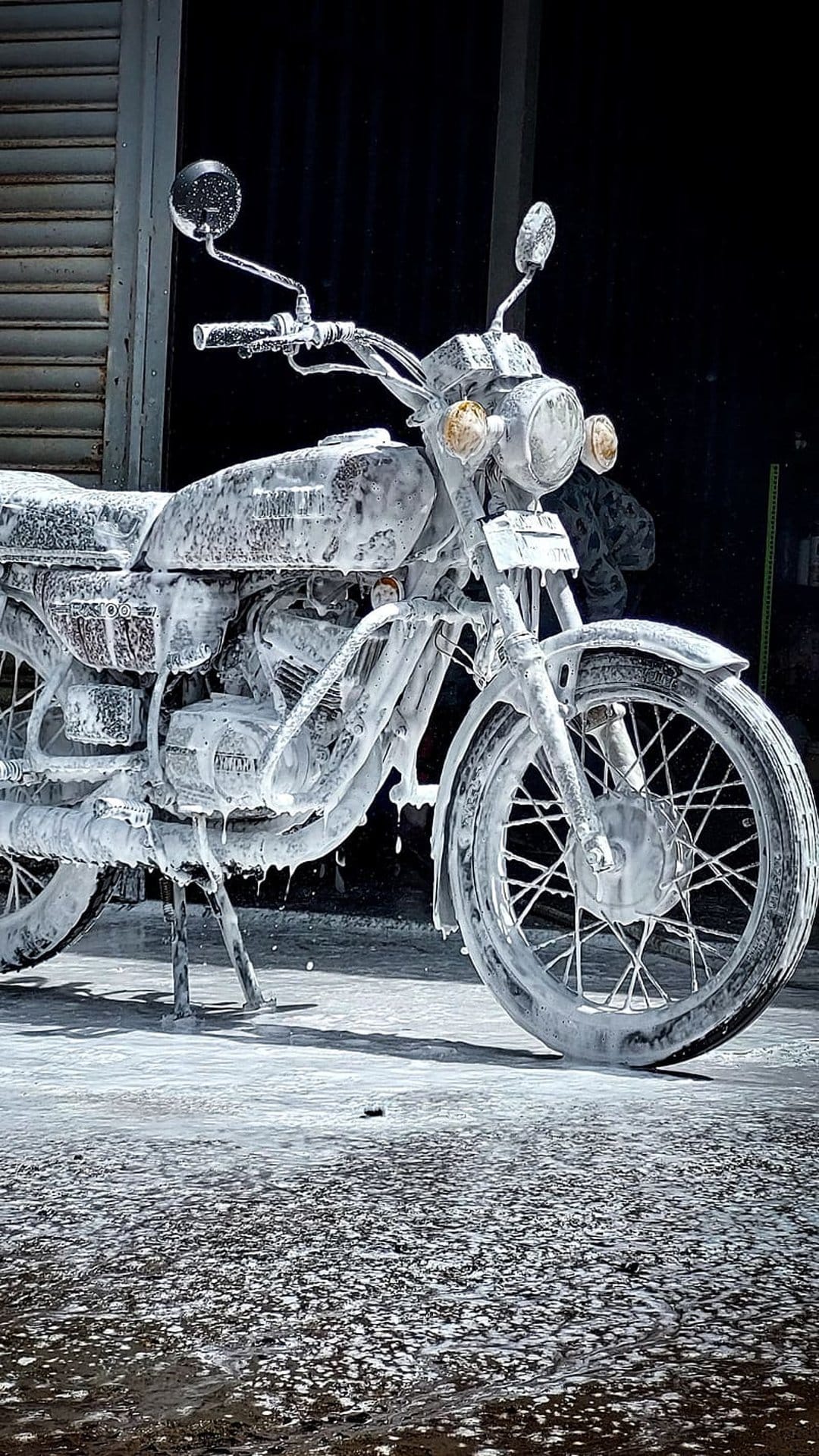 Vintage motorcycle covered in thick white foam suds with visible handlebars and spokes against a dark background.