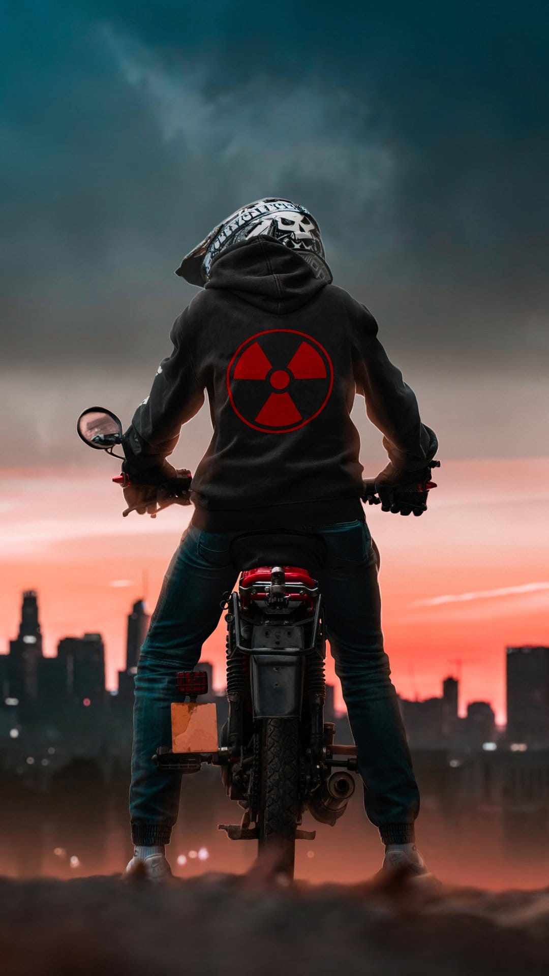 Motorcyclist in a hooded jacket with a red nuclear symbol parked on gravel facing a silhouetted city at sunset.