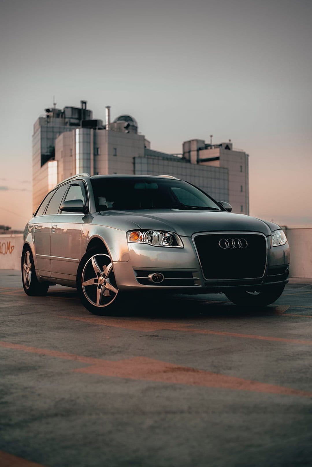 Silver Audi station wagon parked on a concrete rooftop with a tall glass office building under a twilight sky.
