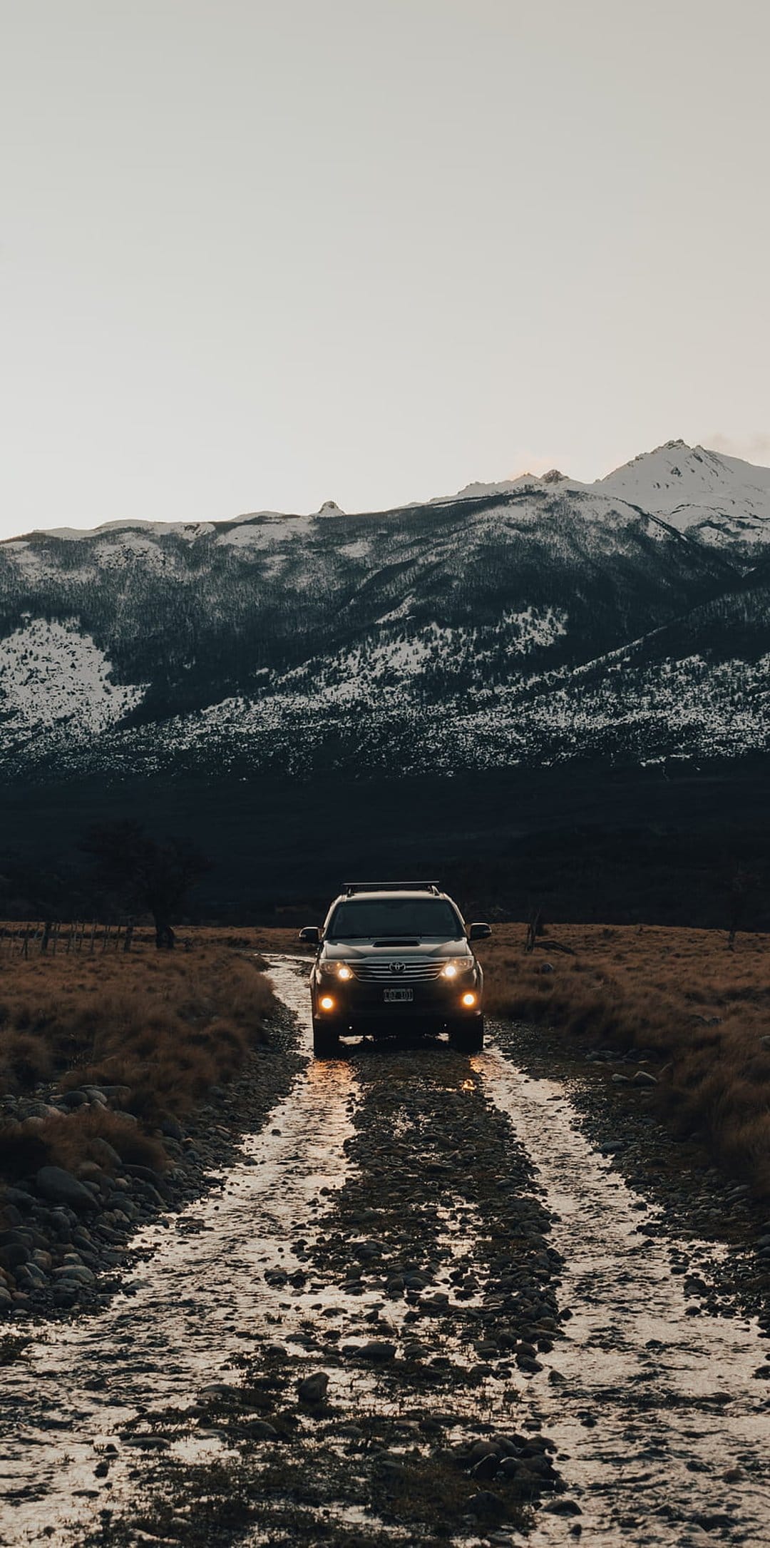 SUV traverses a wet rocky trail toward snowy peaks at twilight with illuminated headlights in a vertical view.