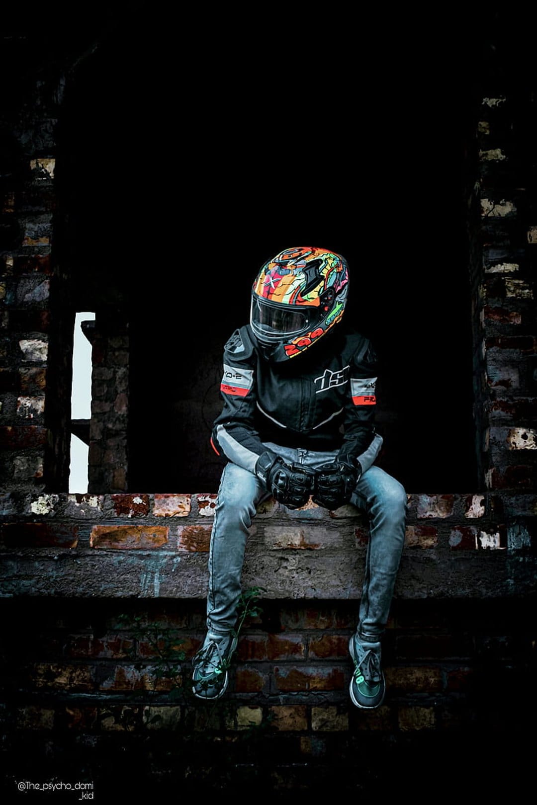 Motorcyclist wearing a multi-colored graffiti helmet sits on a weathered brick wall near a broken window.