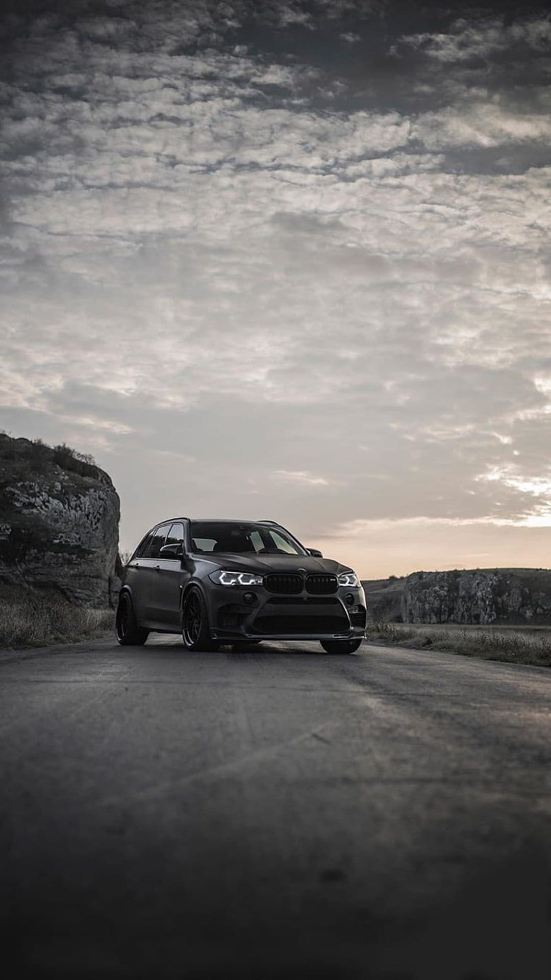 Matte black luxury SUV parked on a mountain road with jagged rocks and dark clouds in a vertical composition.
