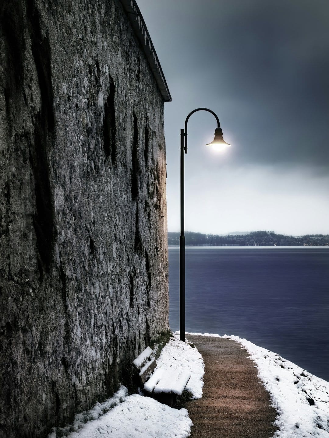 Wrought iron lamppost lighting a snow-covered path next to a stone wall overlooking a dark, churning ocean.