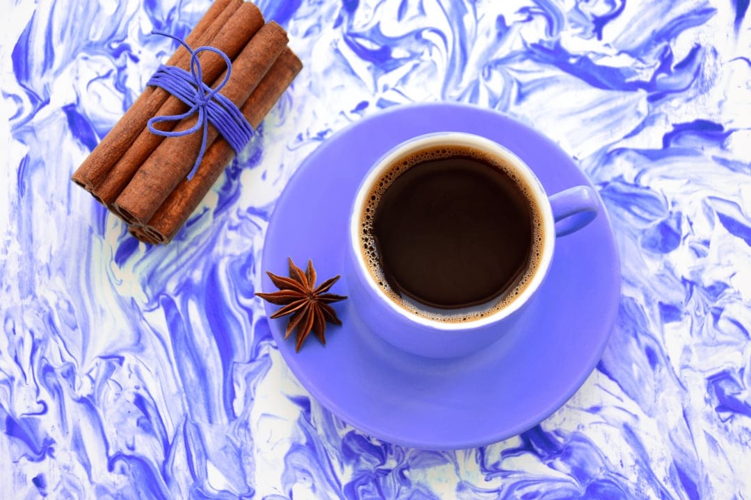 Cup of black coffee on a purple saucer next to cinnamon sticks and star anise over blue marble stone.