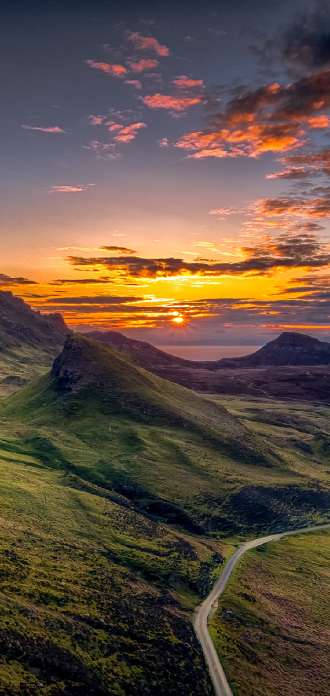 A winding road snakes through green Scottish hills toward a distant loch under a vibrant orange sunset sky.