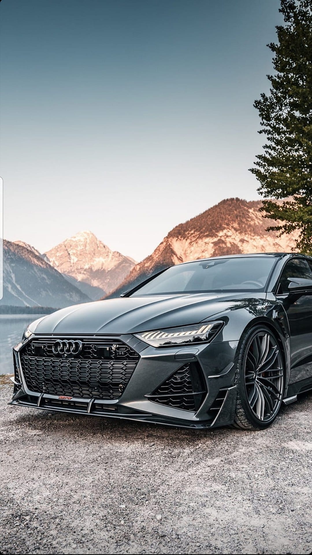 Dark grey Audi RS7 parked on gravel beside a calm lake with sunlit mountain peaks in the background.