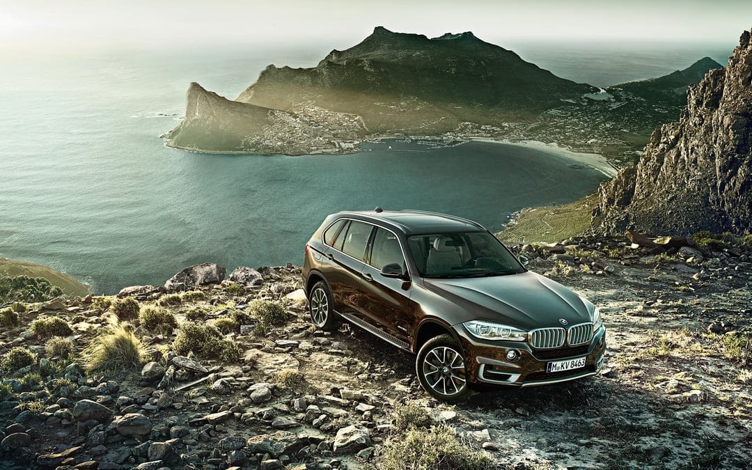 Dark brown BMW SUV on a rocky cliff overlooking a turquoise bay and misty mountains at sunset.