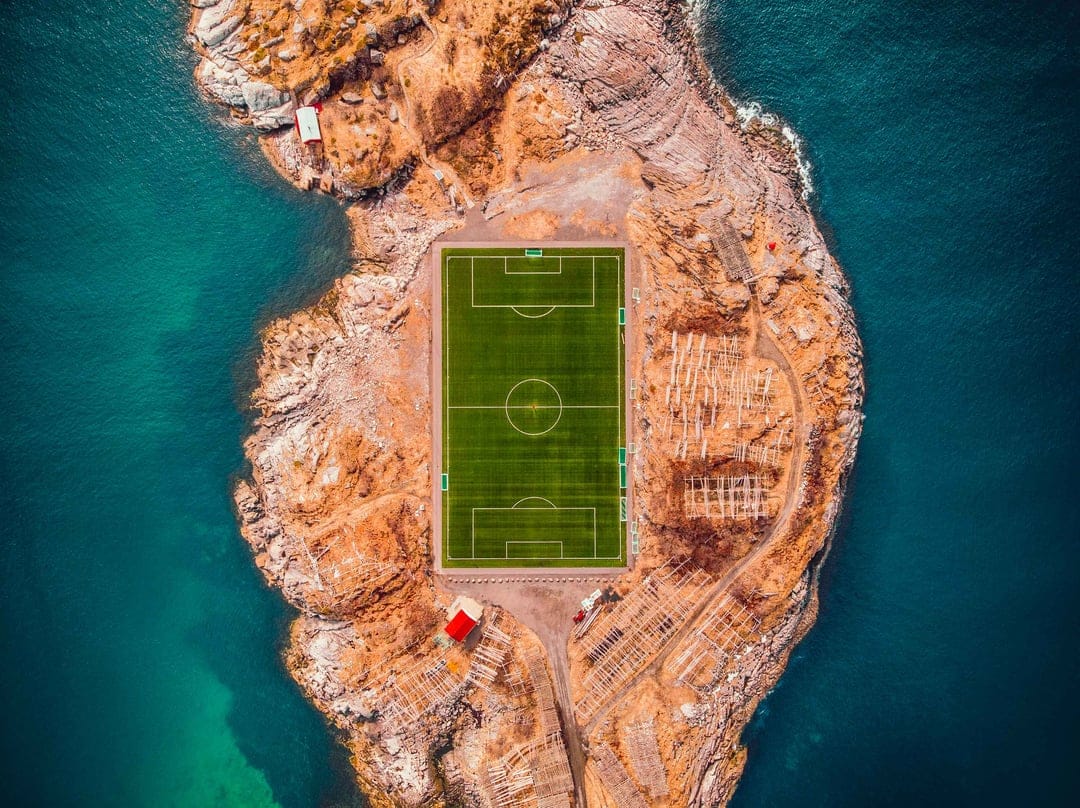 Vibrant green football pitch centered on a jagged rocky island surrounded by swirling turquoise ocean water.