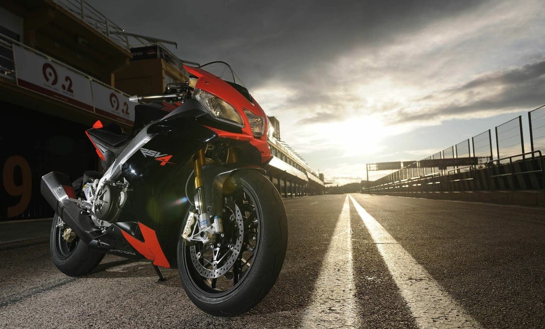 Black and red Aprilia RSV4 motorcycle parked on a sunlit racetrack under a dramatic cloudy sky at dusk.