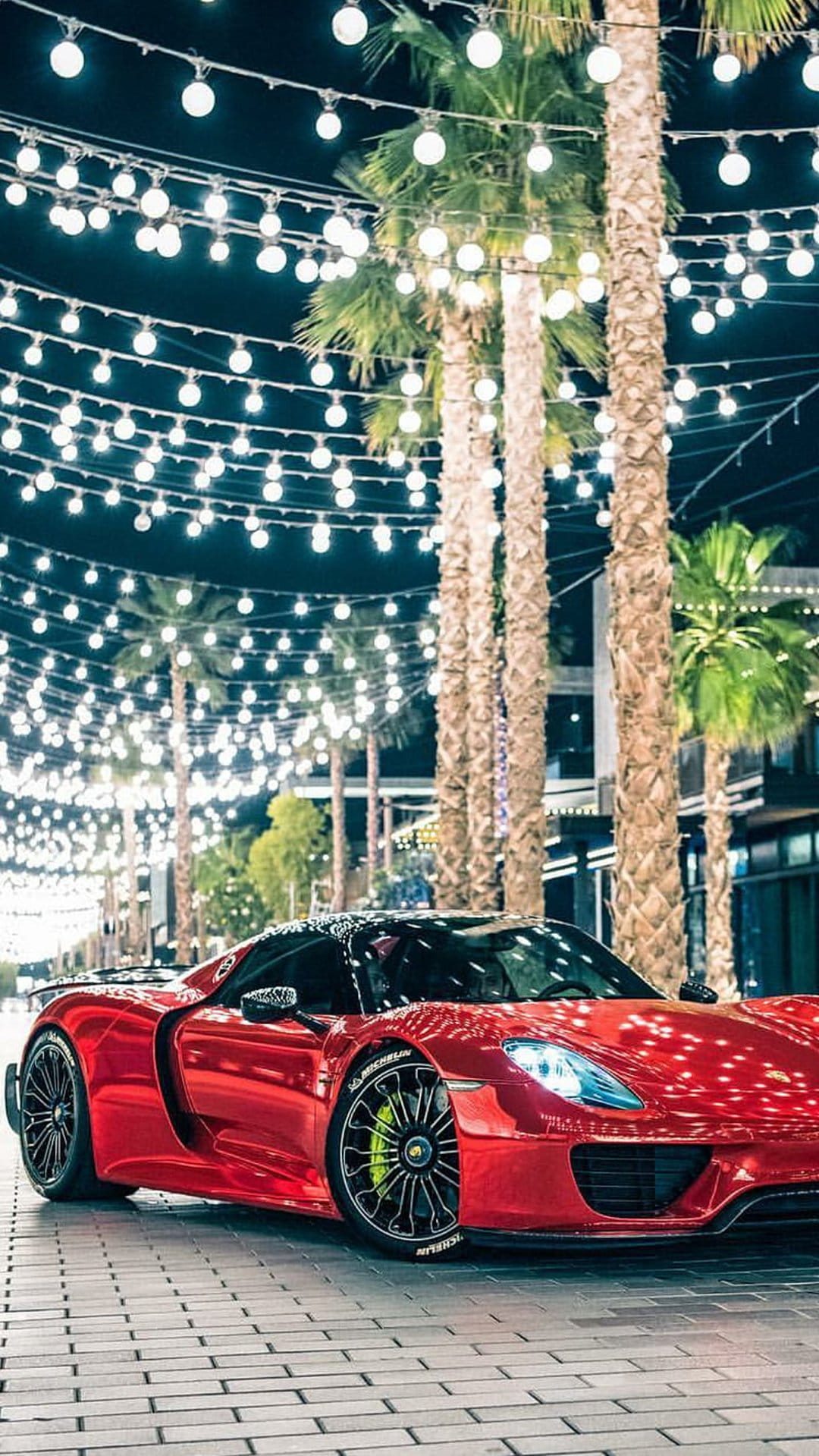 Red sports car parked on a brick path under glowing string lights and tropical palm trees during a summer night.