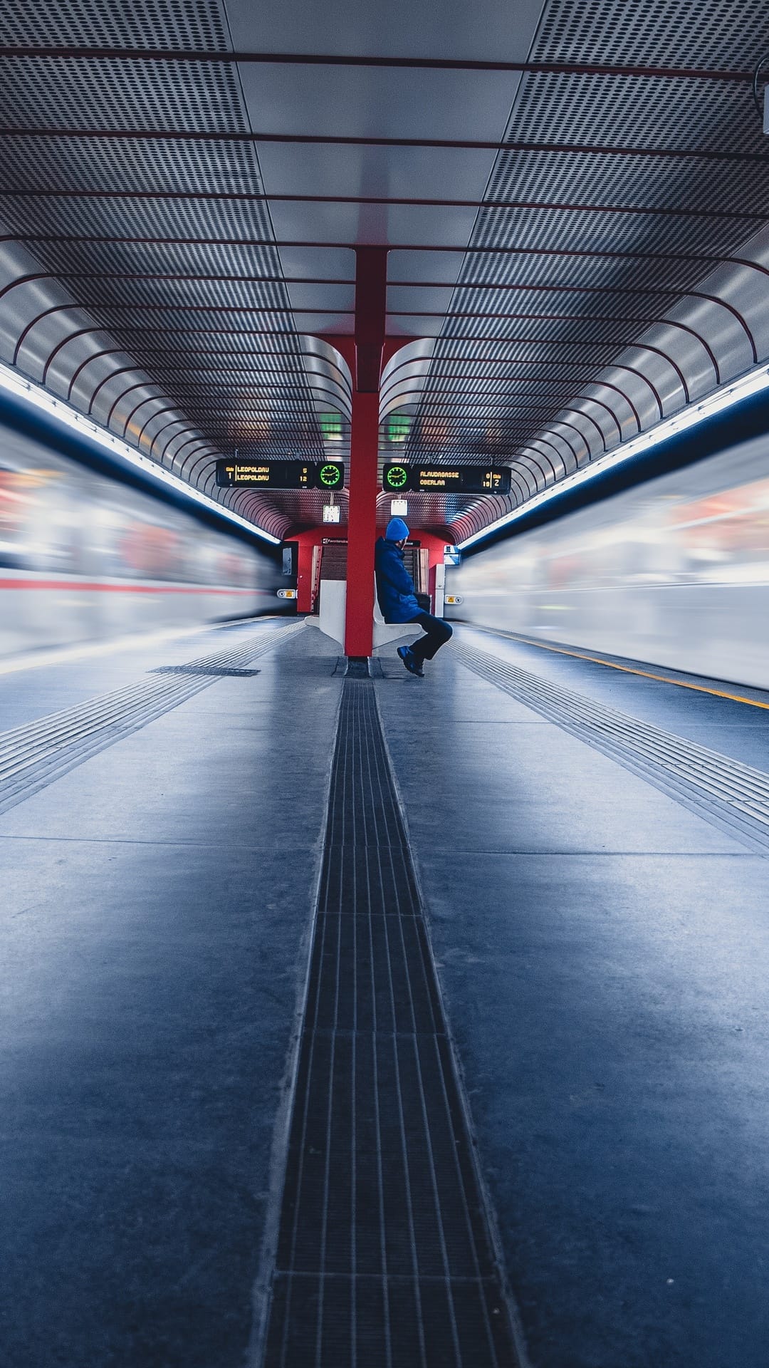 Person in a blue jacket sitting on a subway platform next to long exposure motion streaks of a passing train.