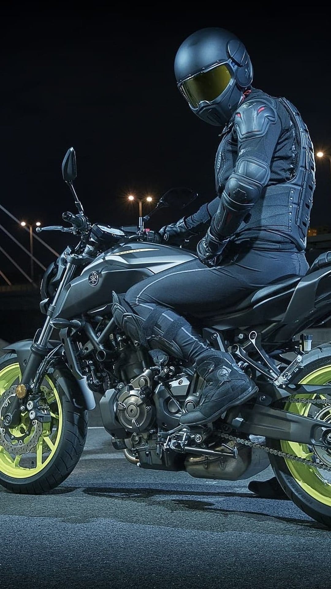 Motorcycle rider on a dark Yamaha bike with neon yellow rims under city streetlights in a vertical night scene.
