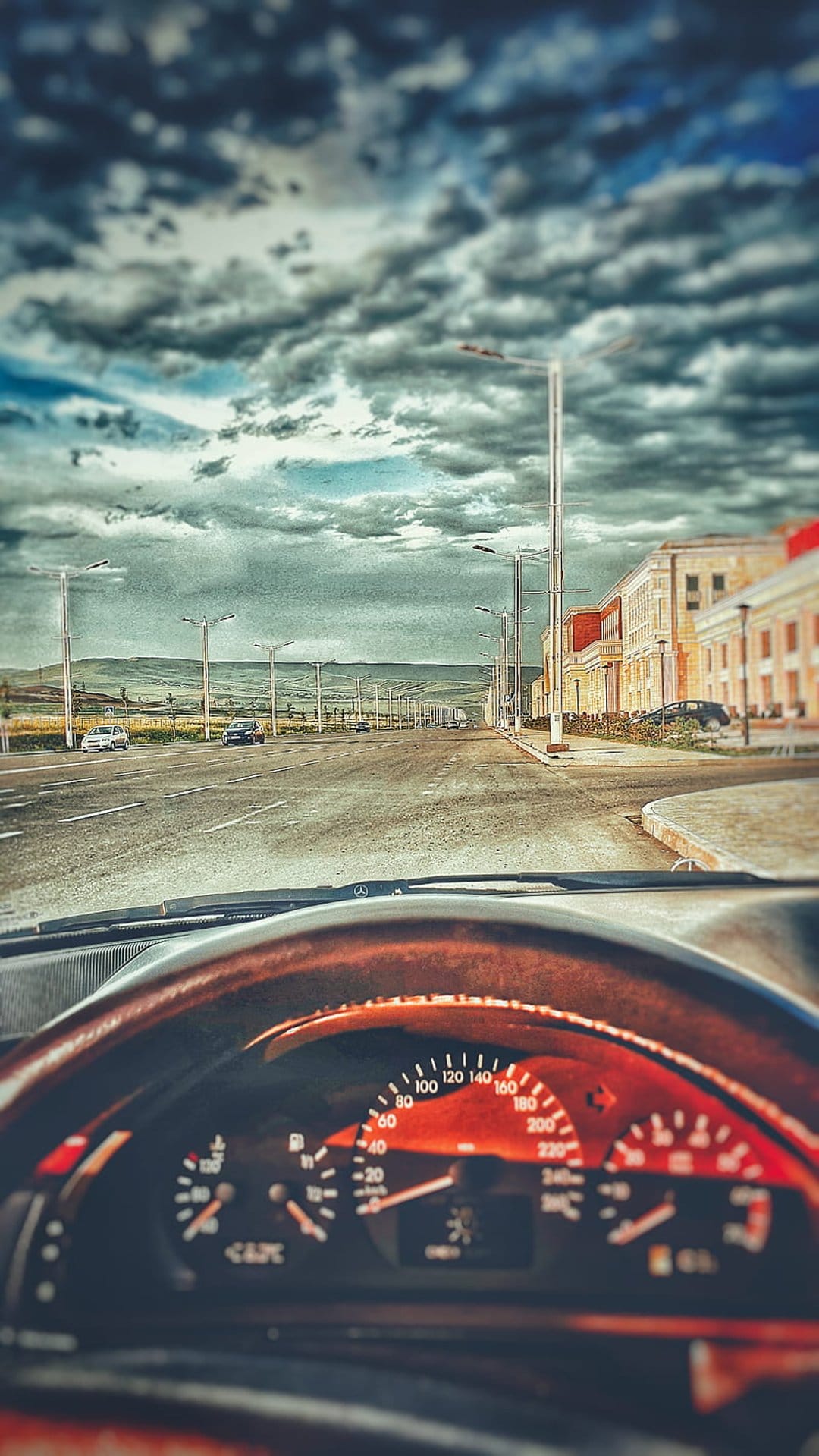 Wooden steering wheel and glowing dashboard gauges seen from the driver's seat on a cloudy urban road trip.