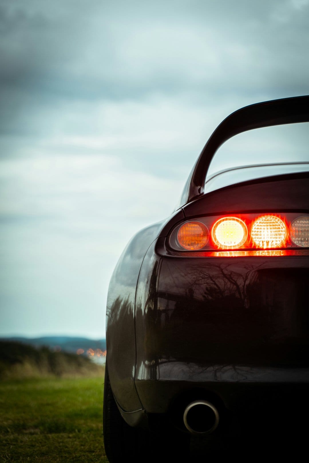Red sports car taillights glow intensely on a dark vehicle with a large spoiler under a cloudy outdoor sky.