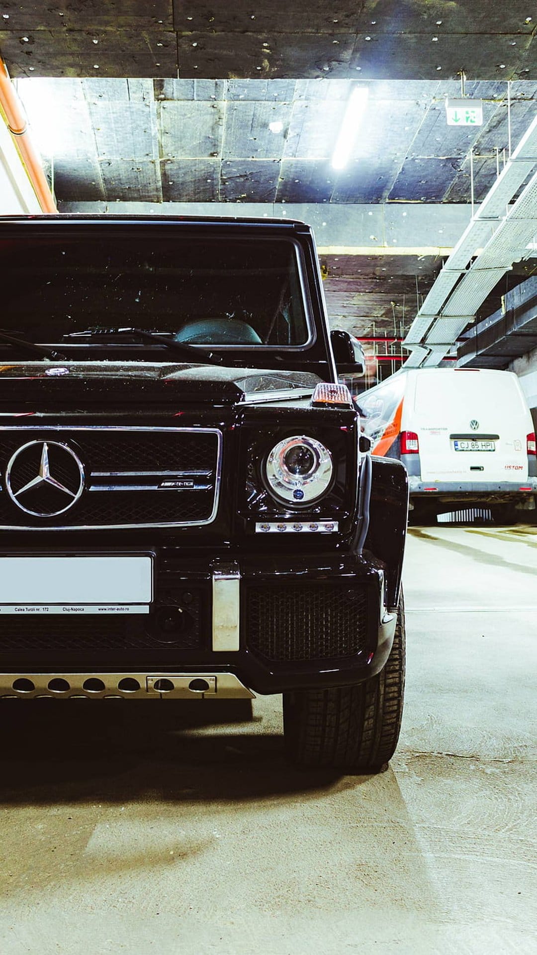 Black Mercedes G-Wagen SUV with a prominent grille parked in a dimly lit garage next to a white service van.