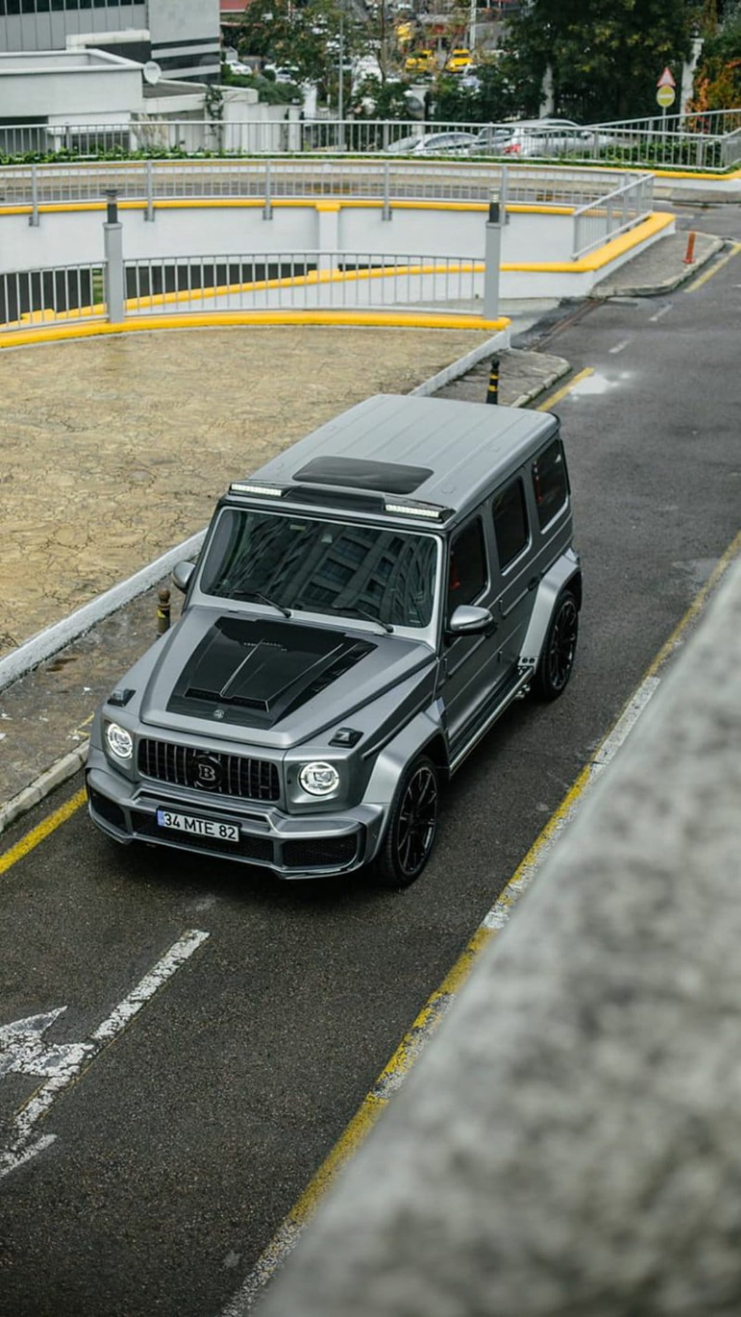 Silver Brabus G-Wagen SUV driving on wet asphalt with yellow lines near a modern urban parking structure.