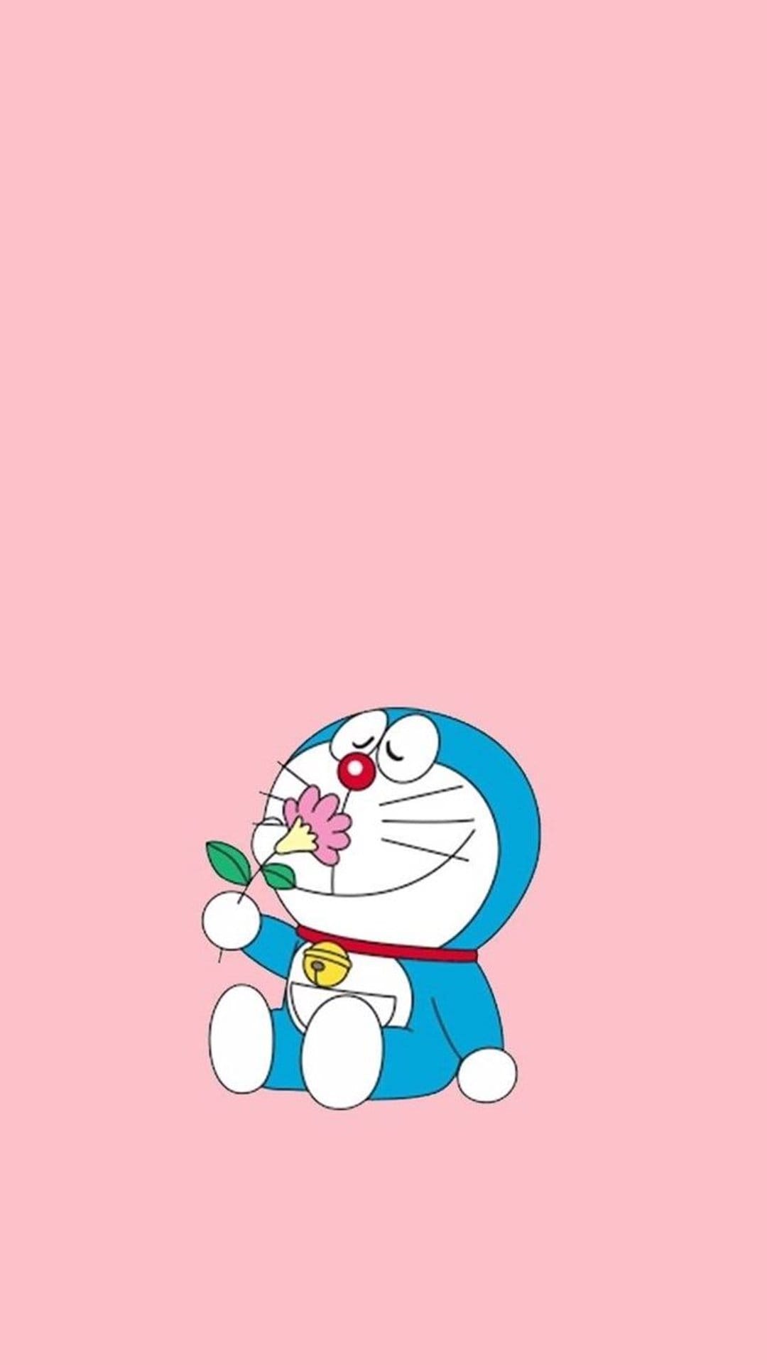 Doraemon smells a small pink flower with closed eyes against a solid light pink background, showing his red nose.
