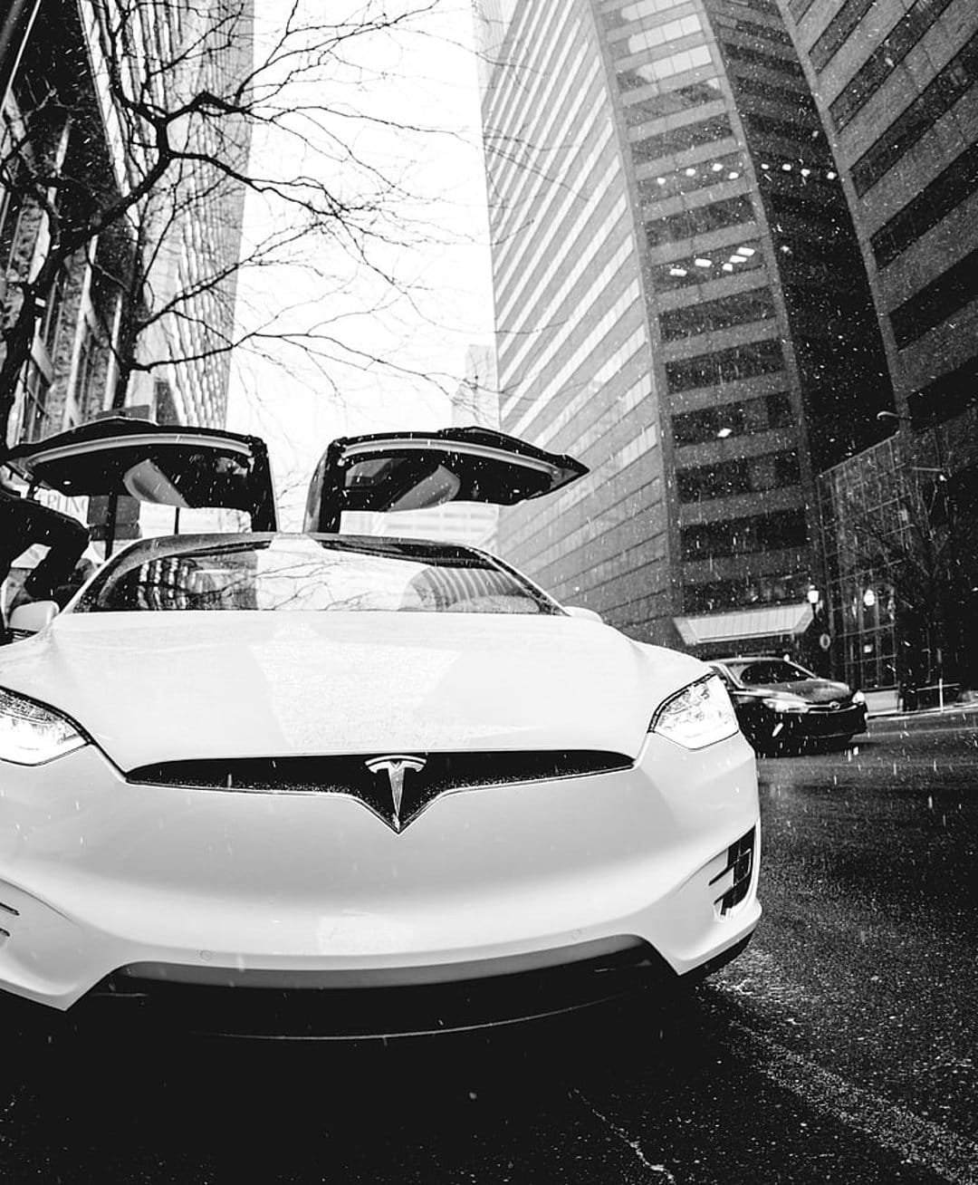 White Tesla Model X with open Falcon Wing doors on a snowy city street framed by tall glass skyscrapers.