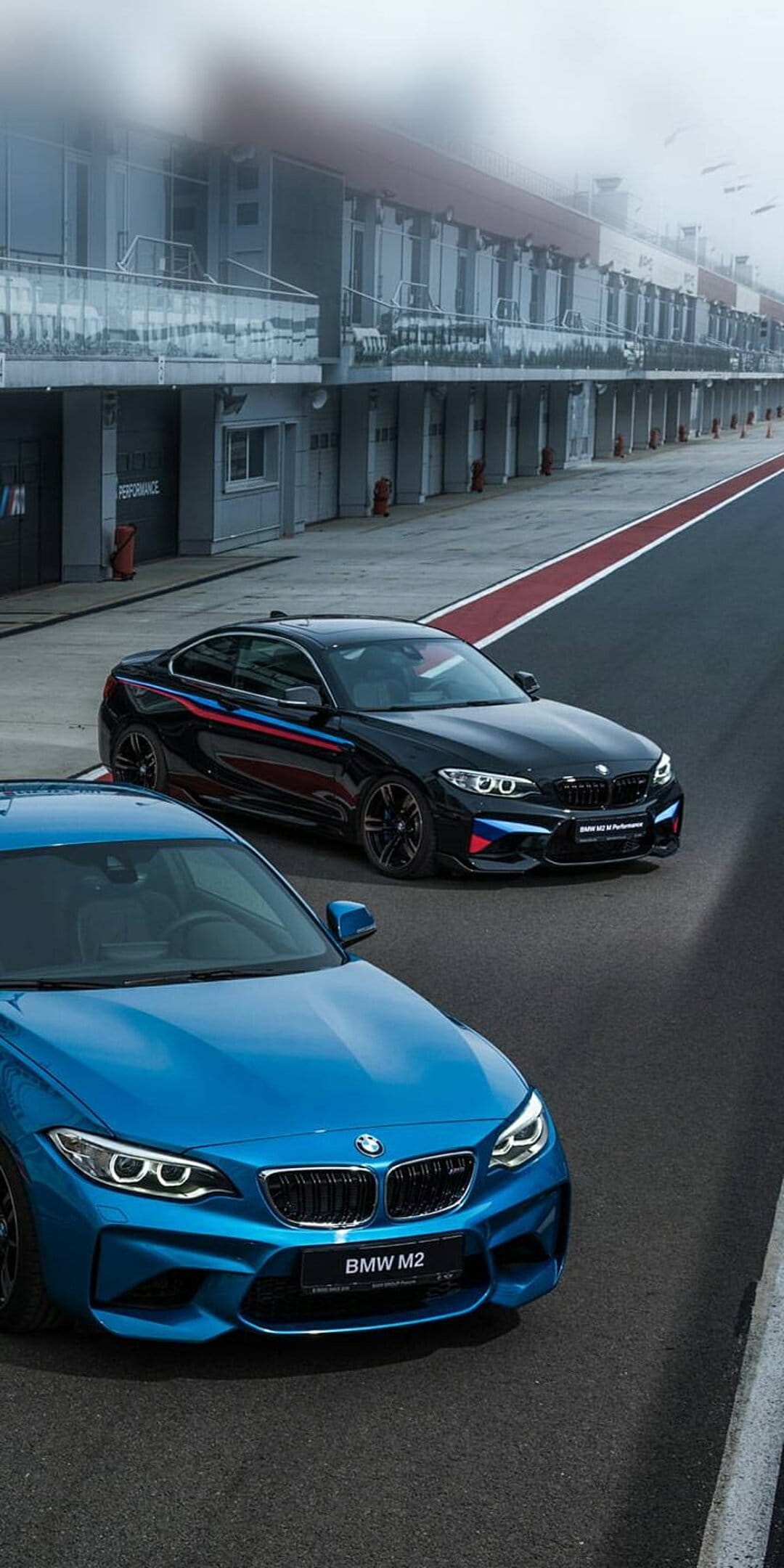 Blue BMW M2 sports car leads a black racing coupe on a misty asphalt track with garage buildings in the distance.
