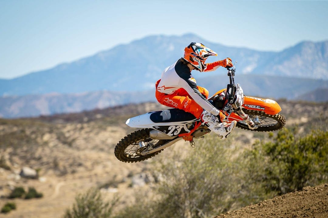Motocross rider in orange gear jumping a KTM dirt bike through the air with blue mountains in the background.