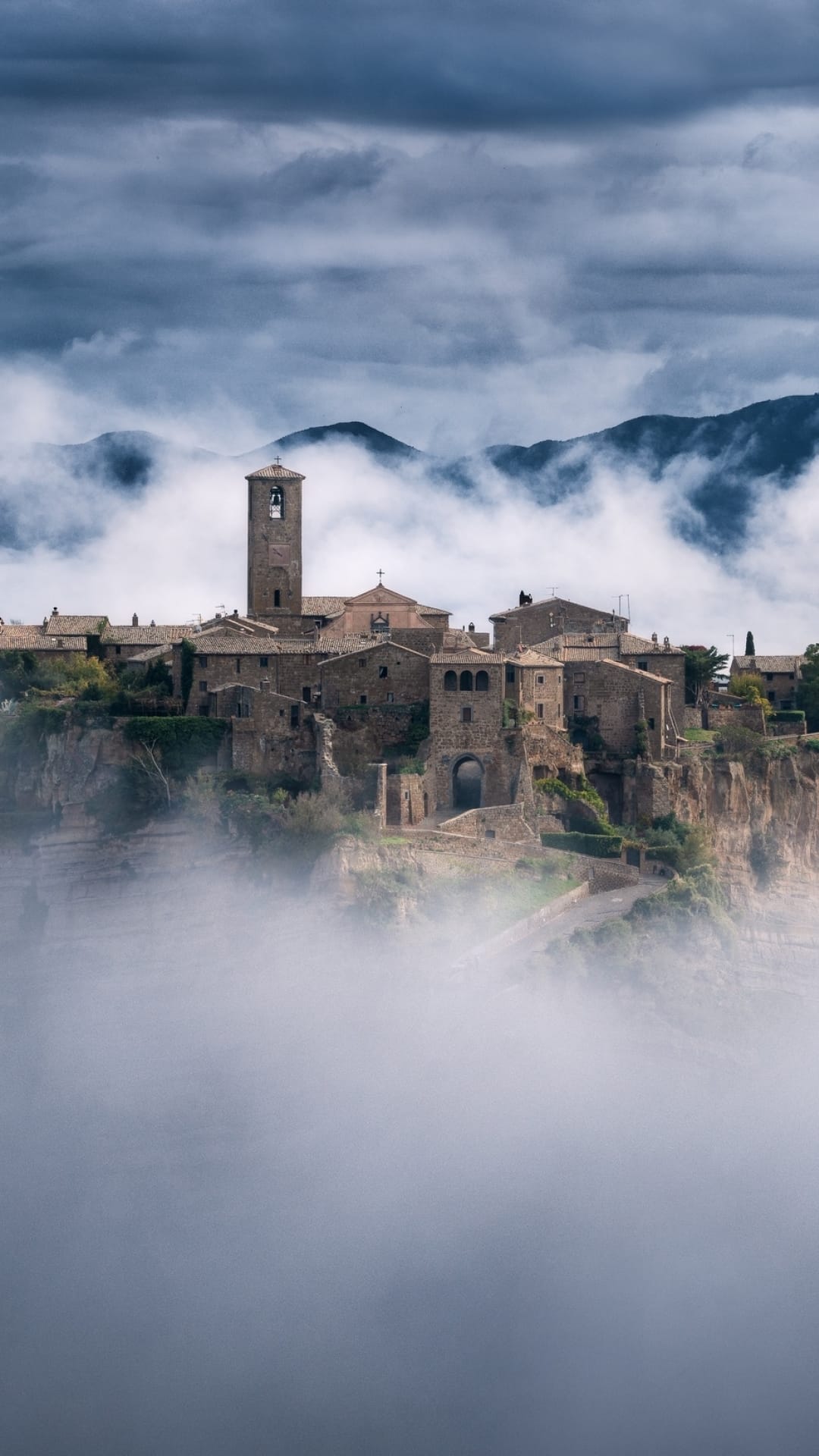 Medieval Italian village with a bell tower rises above thick white clouds under a dramatic gray sky.