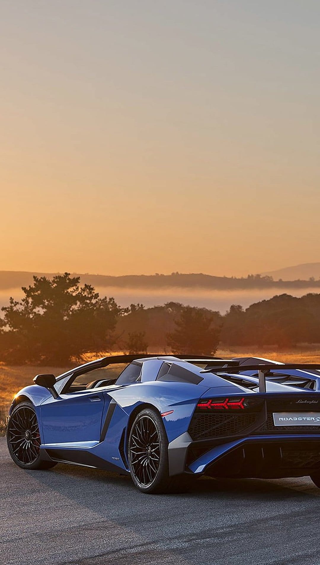 Blue Lamborghini Aventador SV Roadster on a scenic road with a sunrise sky, rolling hills, and lush green trees.