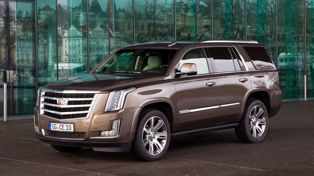 Bronze Cadillac Escalade SUV parked in front of a modern glass building with reflections and chrome wheels.