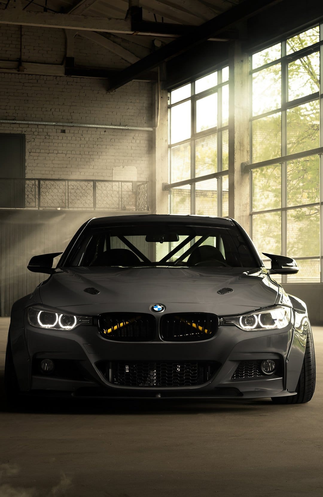 Matte black BMW M3 with a roll cage and widebody kit parked in a sun-drenched industrial warehouse.