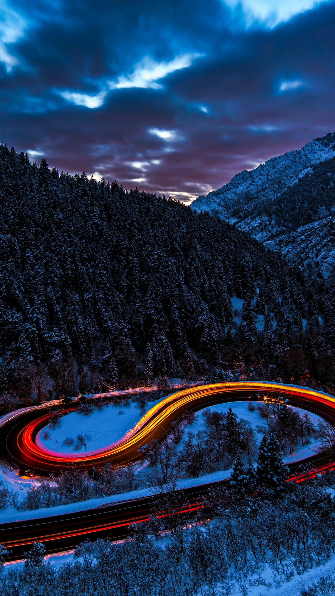 Orange and red light streaks curve along a snow-covered mountain road surrounded by dark evergreen trees.