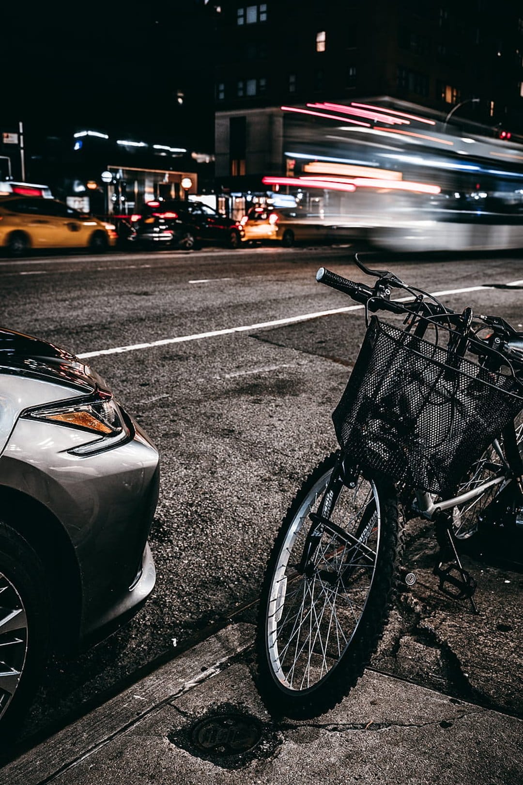 Silver car and bicycle with basket on a wet city street reflecting red and white motion blur light streaks at night.