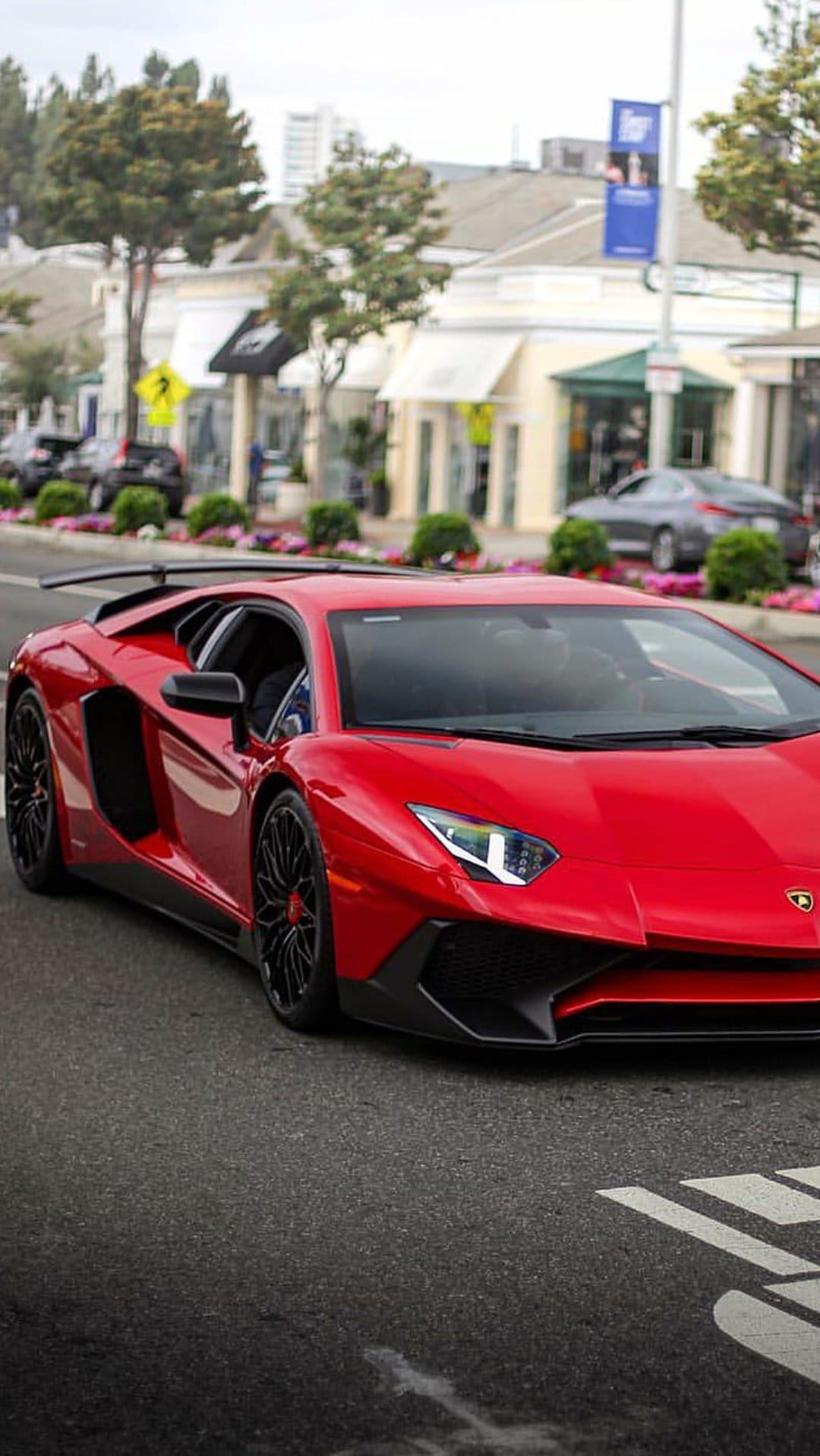Red Lamborghini Aventador driving past shops and green trees on a suburban road with its rear wing visible.