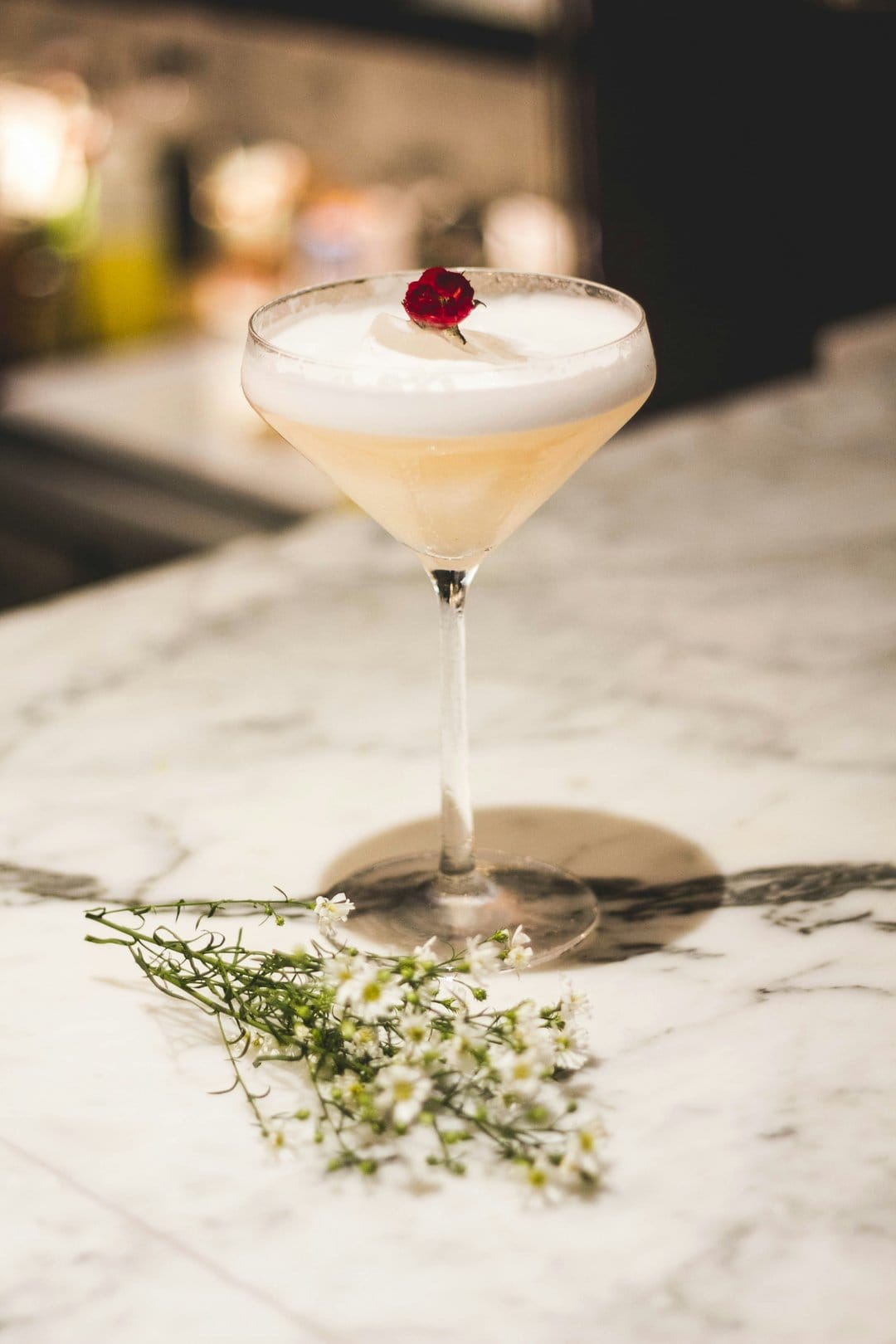 Coupe glass cocktail featuring thick white foam and a red flower garnish resting on a grey marble countertop.