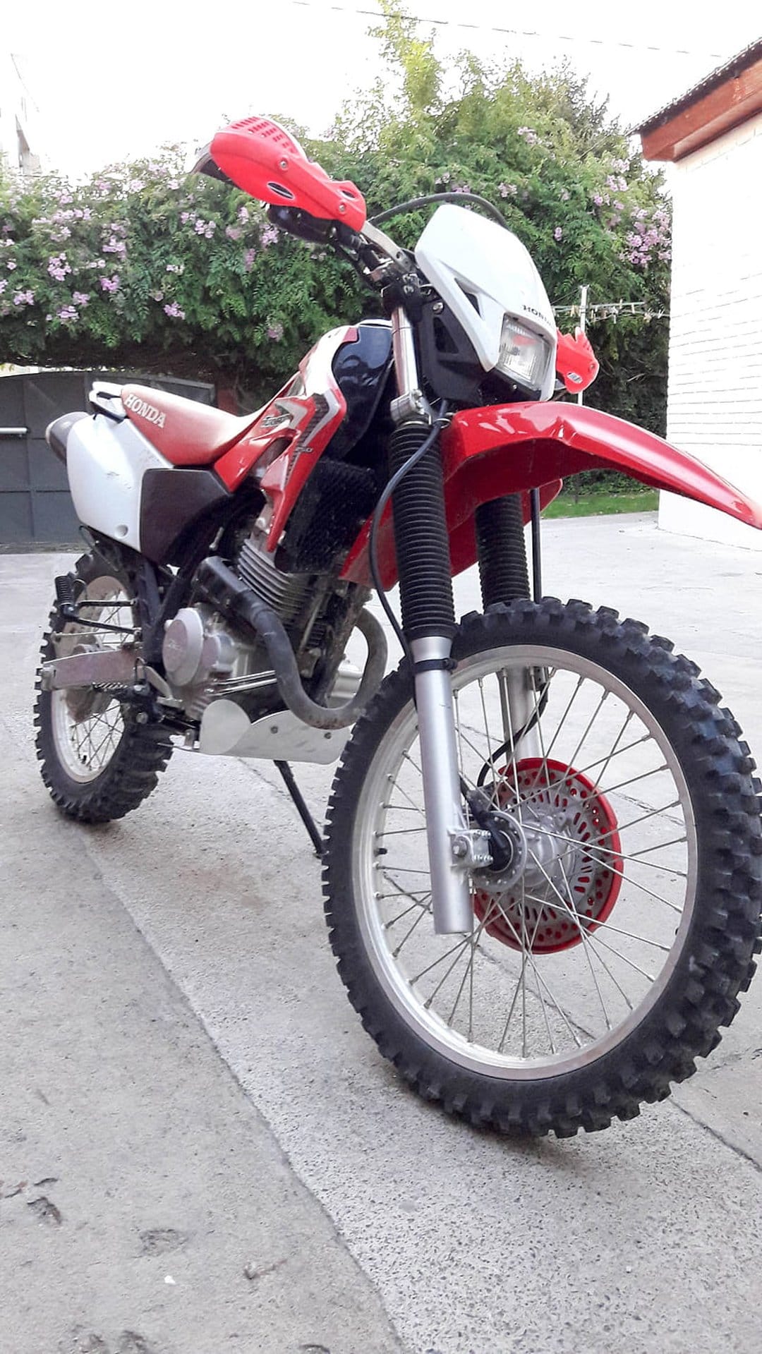 Red Honda dirt bike with knobby tires and white fenders parked on pavement with pink flowers in the background.