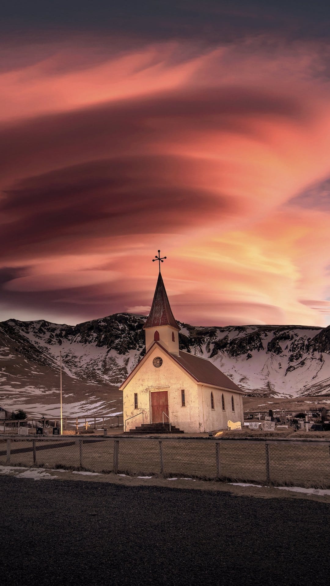 White church with a red spire stands before jagged snow-covered mountains under a dramatic orange and pink sky.