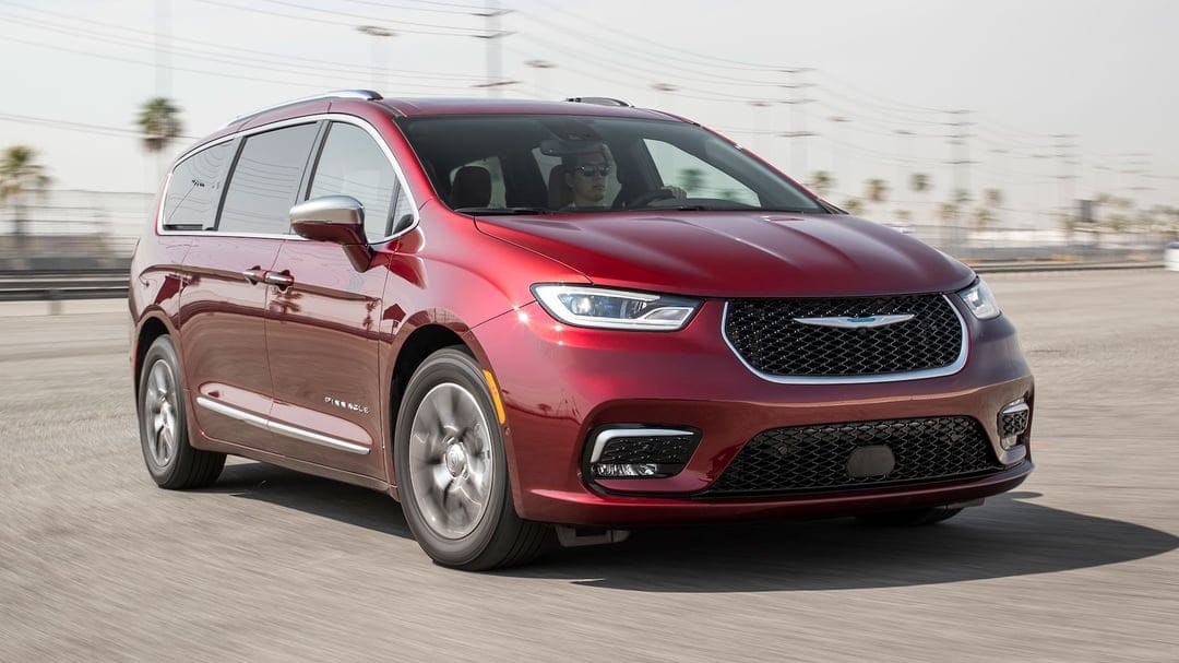 Red Chrysler Pacifica Pinnacle minivan driving on a sunlit paved track with visible motion blur on the wheels.