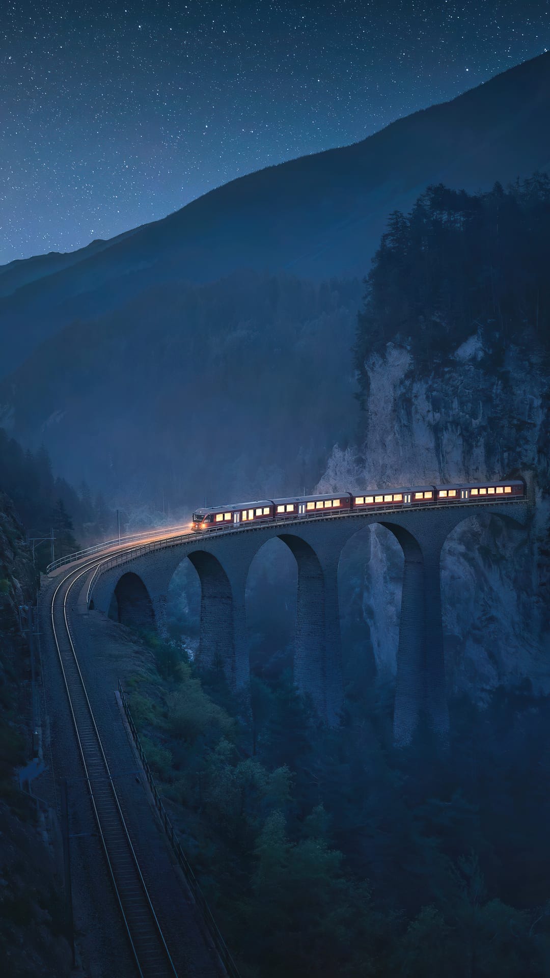 Steam train with lit windows crosses a stone viaduct amidst misty mountains under a starry night sky.