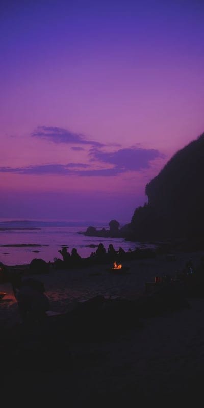 Sunset, twilight, beach, bonfire, purple sky, serene, peaceful, coast, ocean, rocks, silhouettes, evening, warm glow, tranquil, nature, outdoors, relaxation, camping, quiet, colorful.