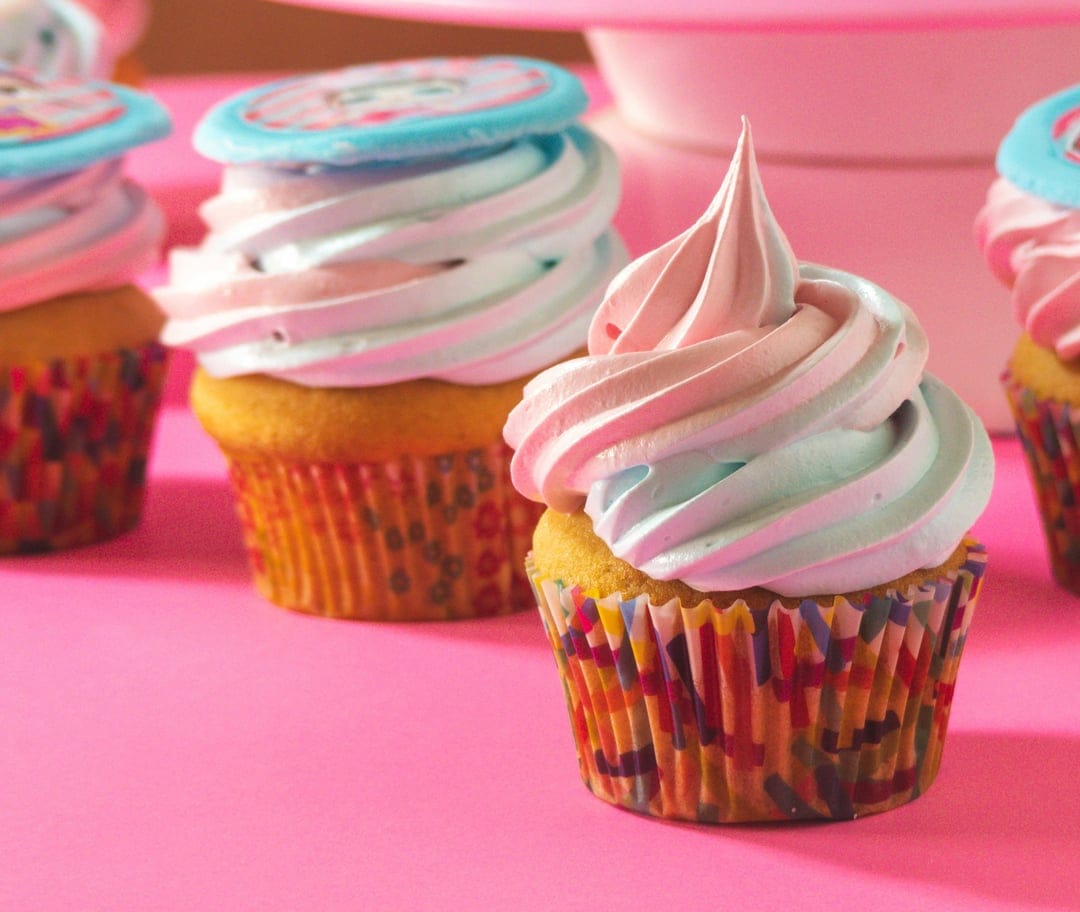 Three cupcakes with pink and blue swirled icing and star toppers sit in confetti wrappers on a pink background.