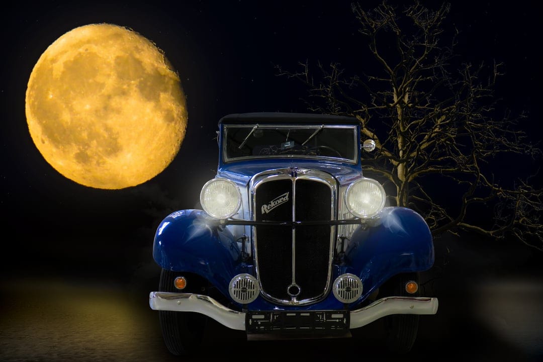 Vintage blue convertible under a full moon with glowing headlights and a leafless tree on a dark night.